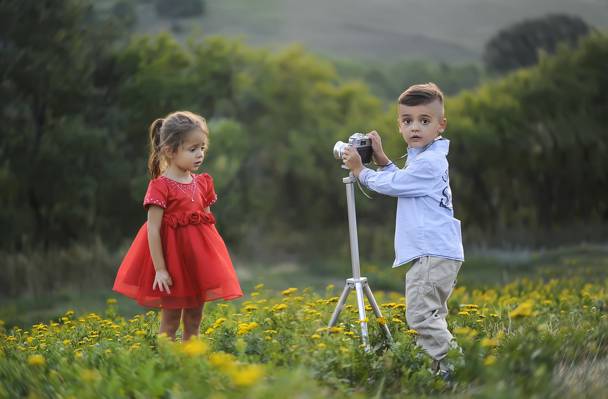 女孩穿着红色的裙子和男孩穿着白色的裙子和裤子拿着相机与三脚架在黄色的花场白天高清壁纸